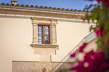A cream building features an ornate window with iron bars, a tiled roof, and a staircase. Blurred pink flowers frame the foreground.