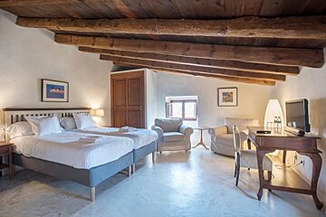 Rustic attic bedroom with twin beds, exposed wooden beams, a cozy sitting area with armchairs, and a desk with a TV.
