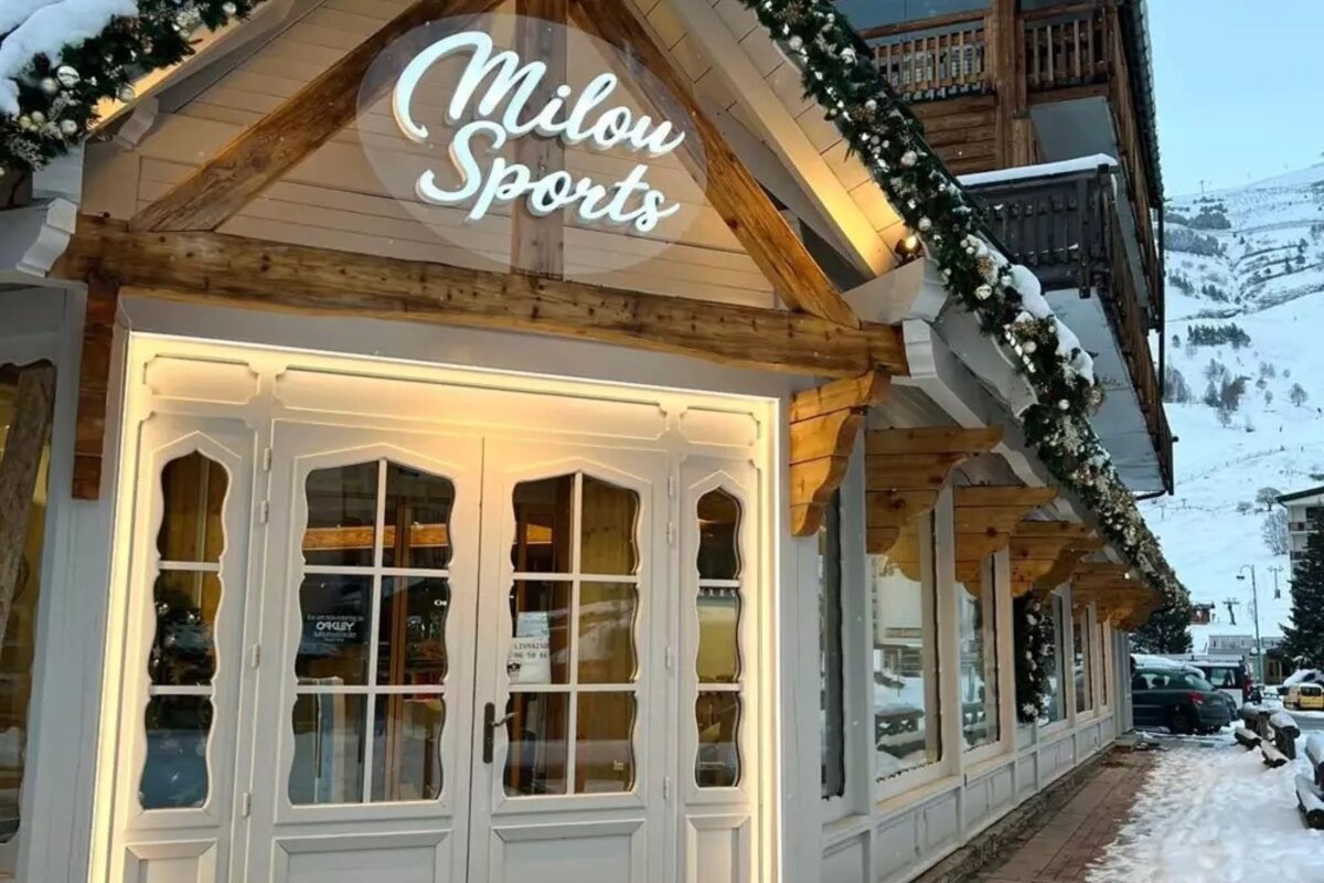 The Milou Sports shop front, adorned with festive garlands and snow, stands in a winter mountain setting with ski slopes visible in the background.