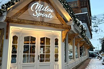 The Milou Sports shop front, adorned with festive garlands and snow, stands in a winter mountain setting with ski slopes visible in the background.