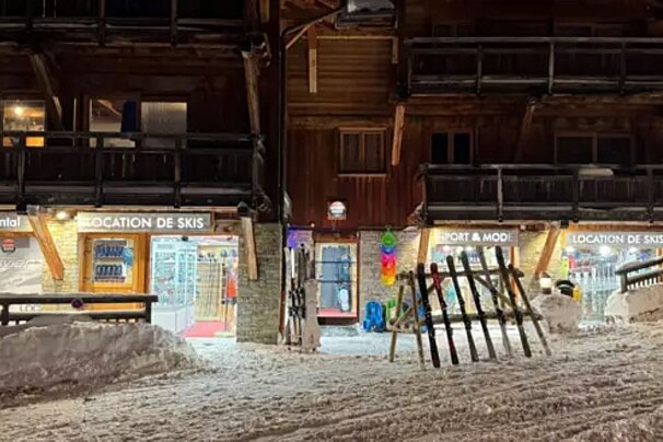 Snowy night scene outside wooden ski rental shops. Illuminated storefronts, with skis propped up and snow covering the ground.
