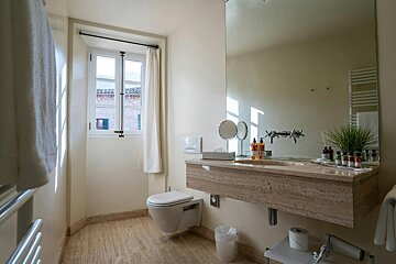 A bright, elegant bathroom featuring travertine flooring, a wall-mounted toilet, a floating stone vanity, and a large mirror by a sunlit window.