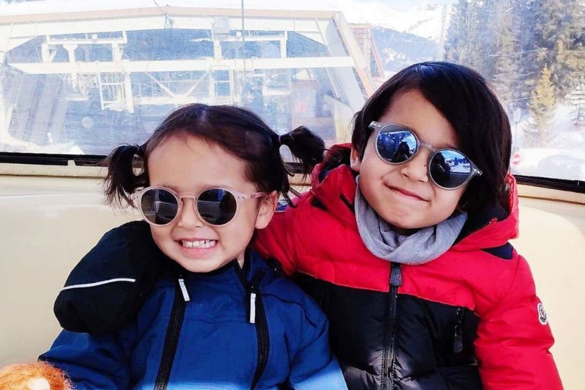 Two happy kids in winter jackets and sunglasses smile from inside a gondola, with a snowy mountain landscape visible outside.