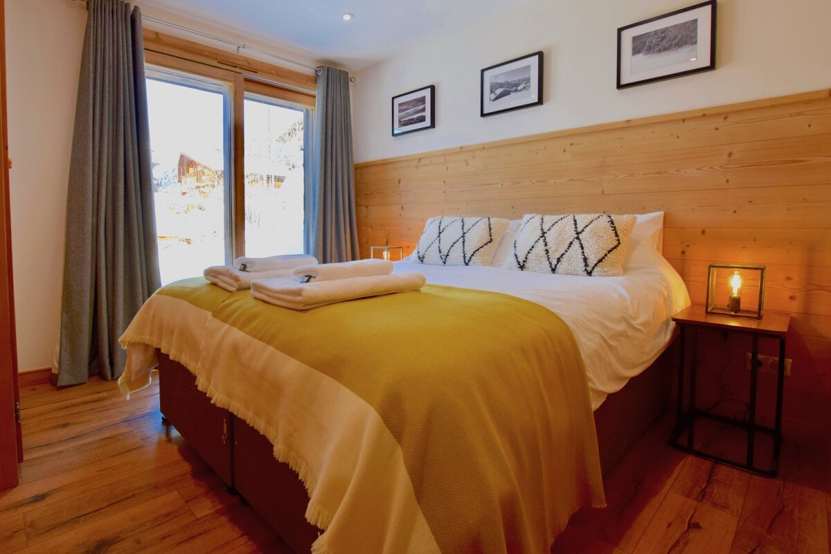 A cozy bedroom with a large window showcasing snow-covered scenery. It features a wooden headboard, yellow and white bedding, and a glowing bedside lamp.