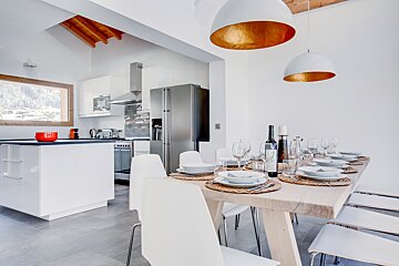A modern open-plan kitchen and dining area with a set wooden table, white chairs, and large windows revealing a snowy mountain landscape.