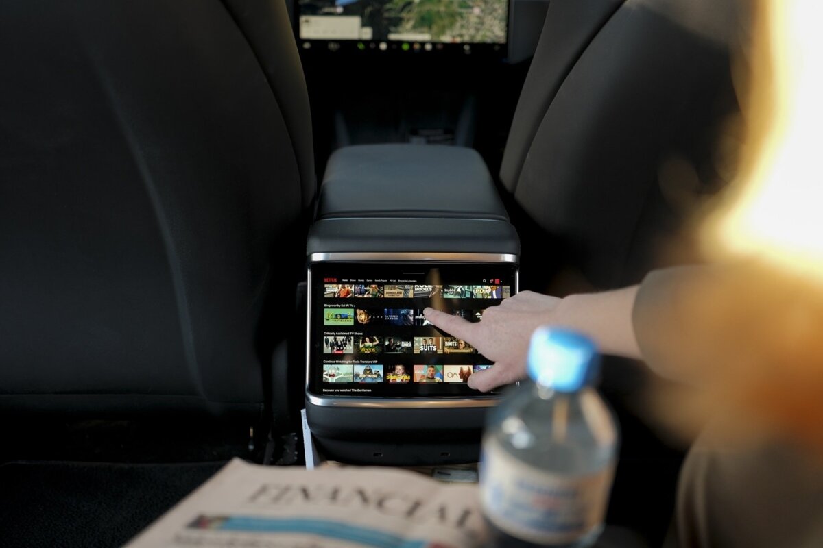 A hand interacts with a Netflix-playing screen mounted on a car's rear center console. A water bottle and newspaper are visible in the foreground.