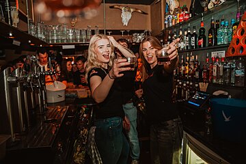 Two women holding up shot glasses in front of a bar full of liquor
