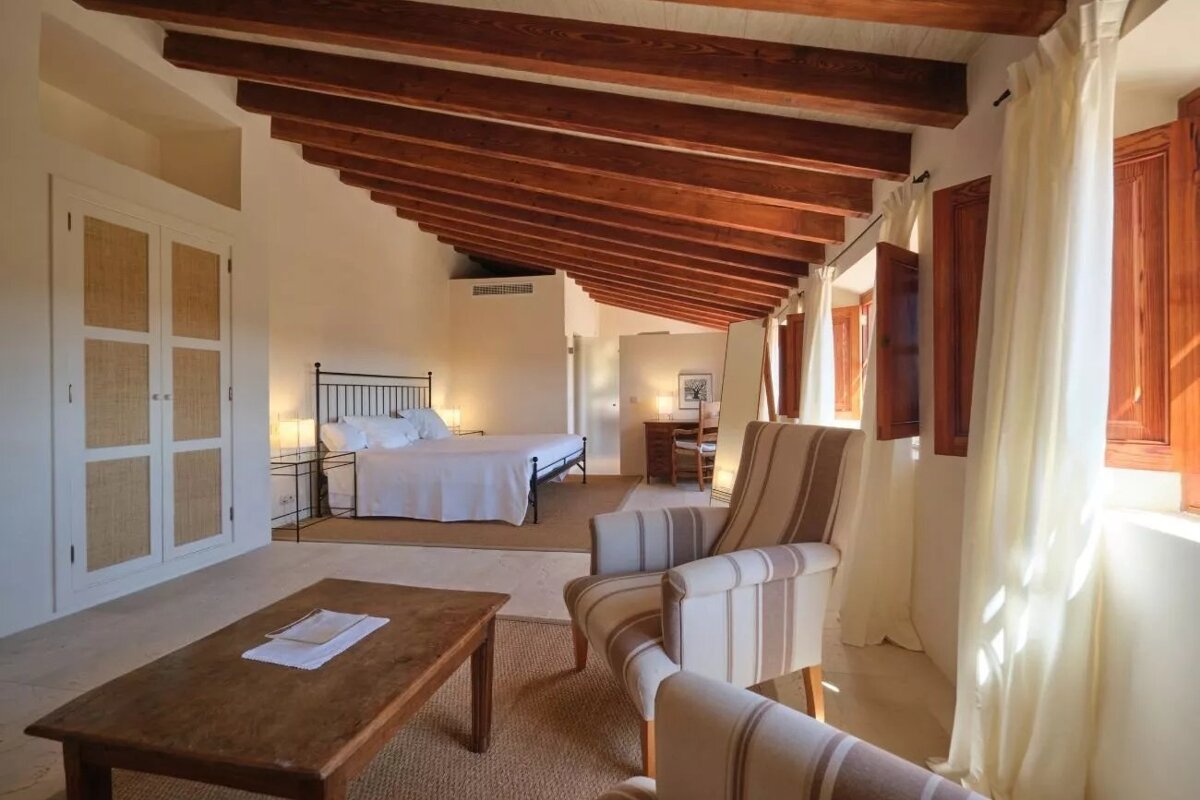 A rustic bedroom with a vaulted wooden beam ceiling, white walls, a large bed, and a sitting area with striped armchairs and wooden window shutters.