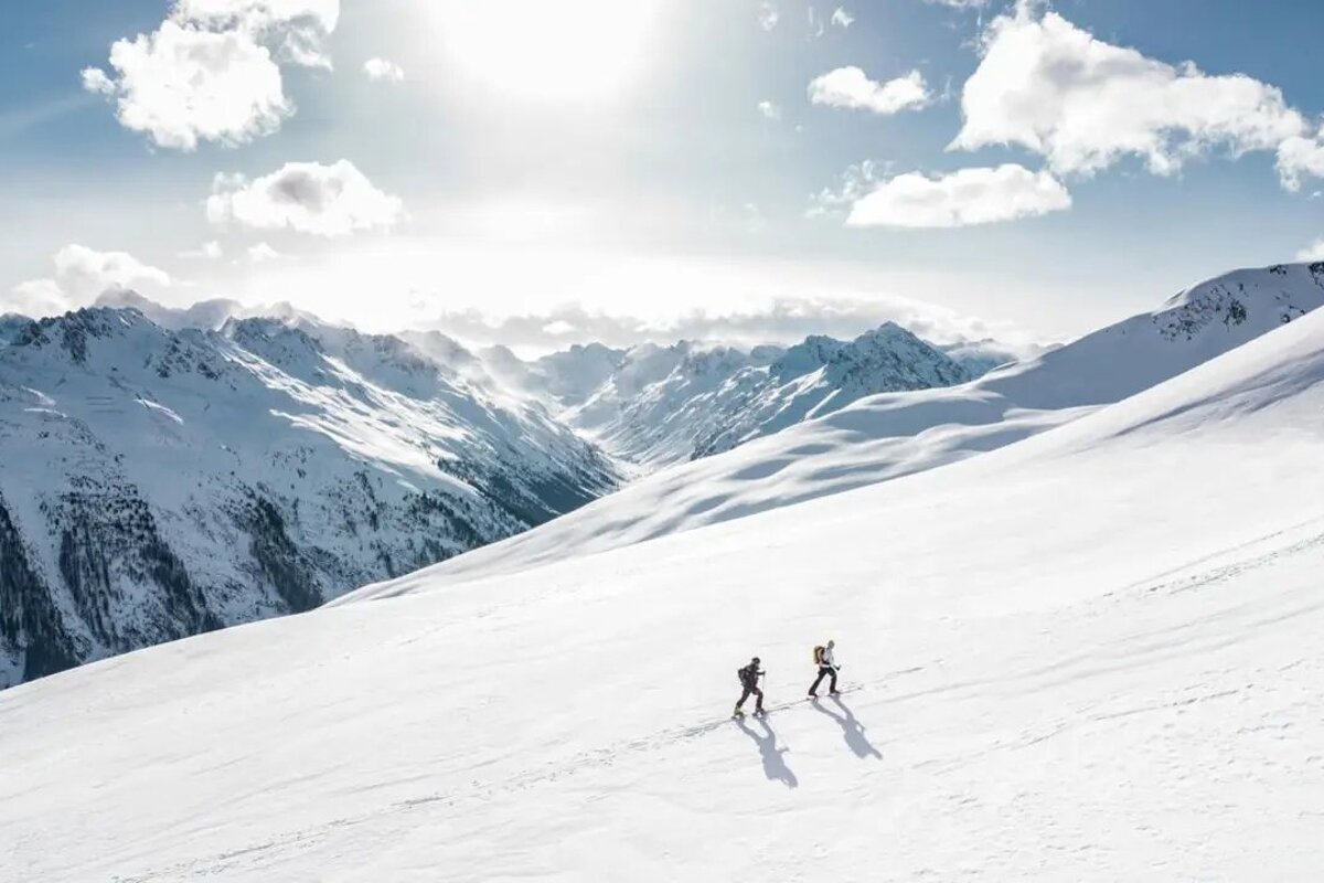 Two ski tourers traverse a vast, snow-covered mountain slope under a bright, sunny sky. Majestic peaks line the distant horizon.