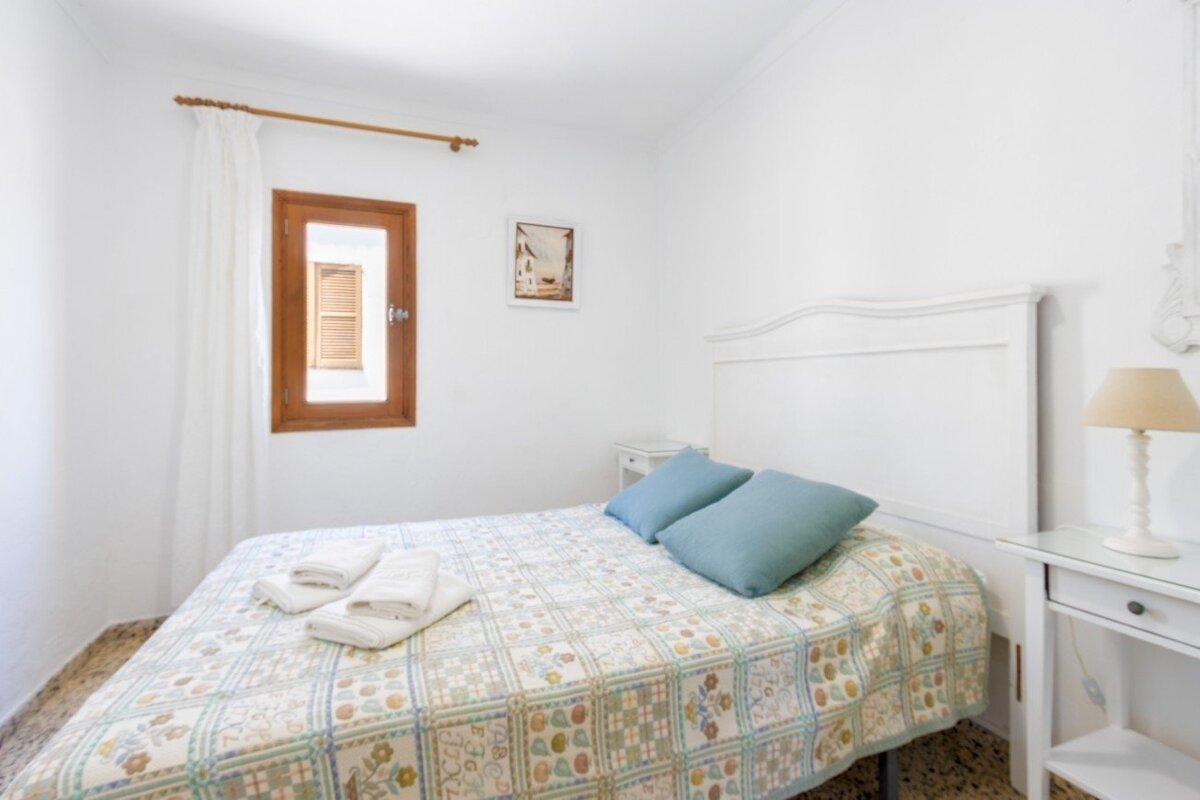 Bright white bedroom with a bed, patterned quilt, blue pillows, wooden-framed window, and a bedside lamp on a white nightstand.
