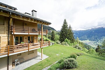 A rustic wooden chalet with balconies on a green hillside, overlooking a lush valley and distant snow-capped mountains under a cloudy sky.