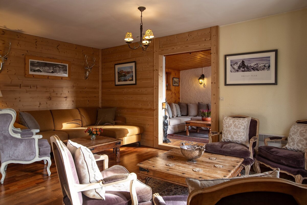 A cozy, wood-paneled living room with a large sofa, ornate armchairs, wooden coffee tables, warm lighting, and mountain decor with deer heads.