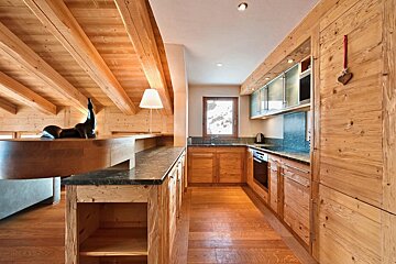 A rustic-modern kitchen featuring extensive light wood cabinetry, floors, and exposed ceiling beams. An L-shaped counter, appliances, and a window with a snowy backdrop.