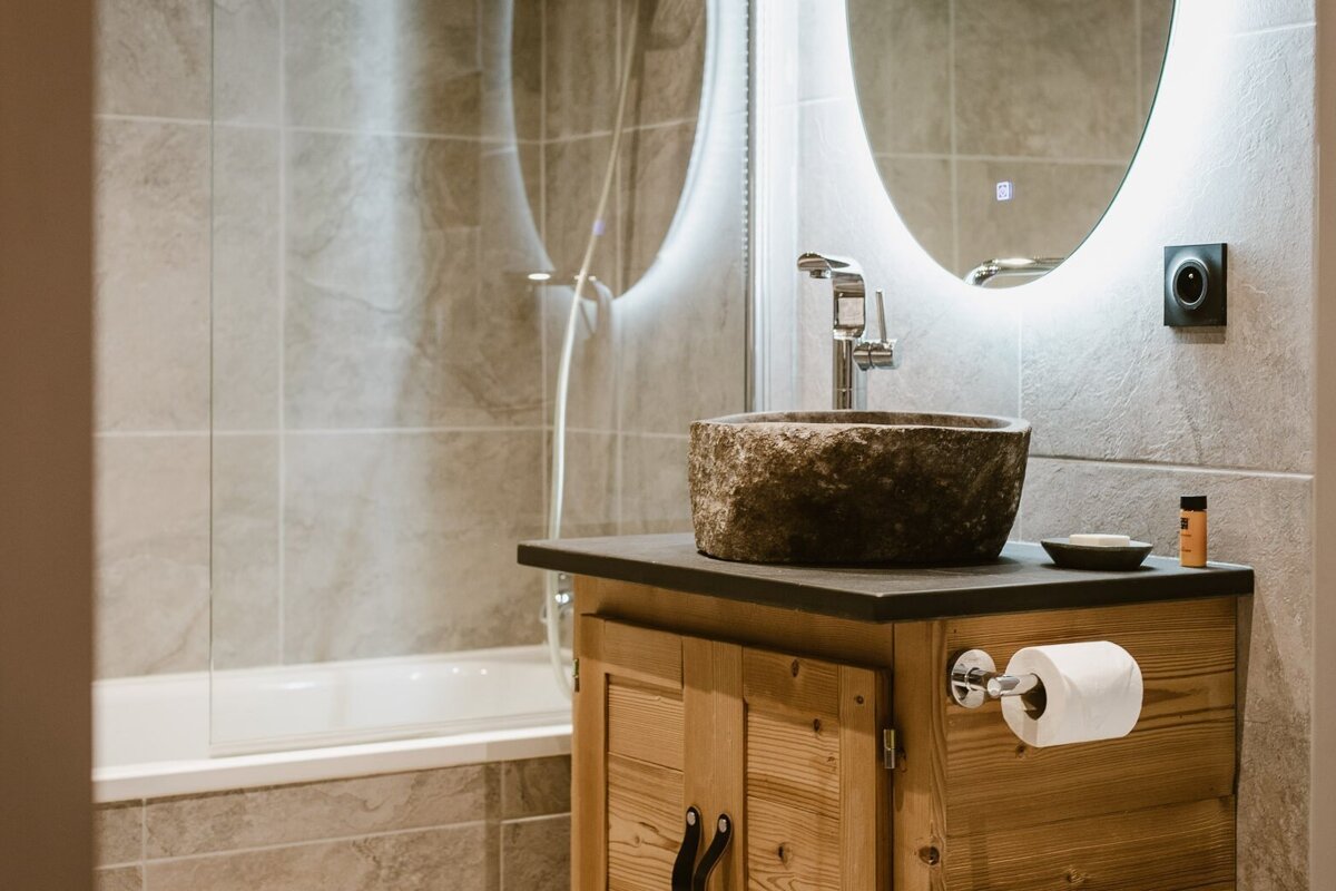 A modern bathroom features a rustic stone sink on a wooden vanity with a lighted mirror, beside a tiled bathtub/shower.