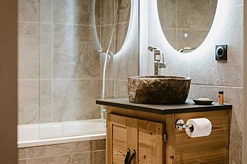 A modern bathroom features a rustic stone sink on a wooden vanity with a lighted mirror, beside a tiled bathtub/shower.