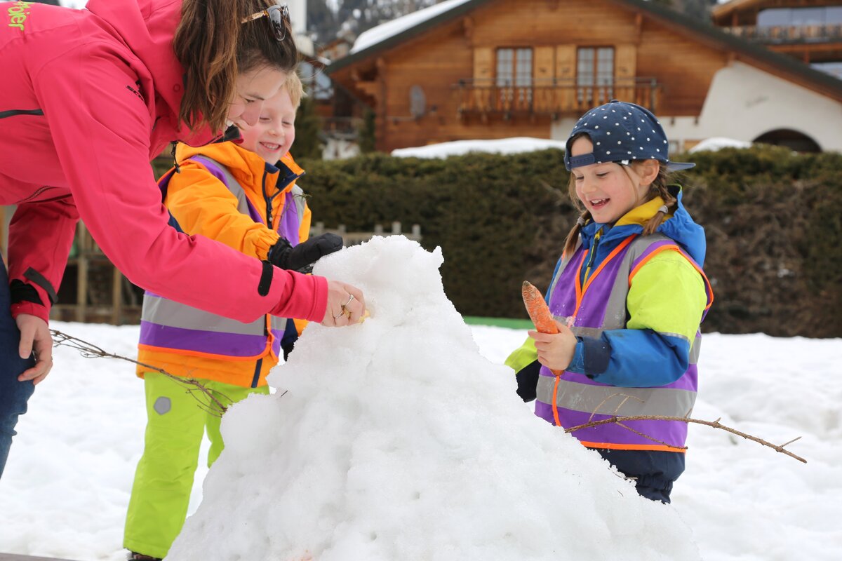 An adult and two smiling children are building a snowman in the snow. One child holds a carrot, while they enjoy the snowy outdoor activity.