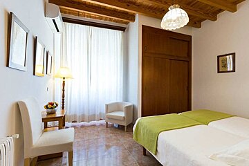 A well-lit bedroom features twin beds with green throws, a beamed ceiling, large window with white curtains, tiled floor, and a wooden wardrobe.