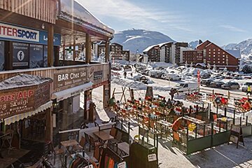 A bustling snowy mountain resort scene with shops, restaurants, outdoor seating, parked cars, and people, all under a bright sky with mountains.