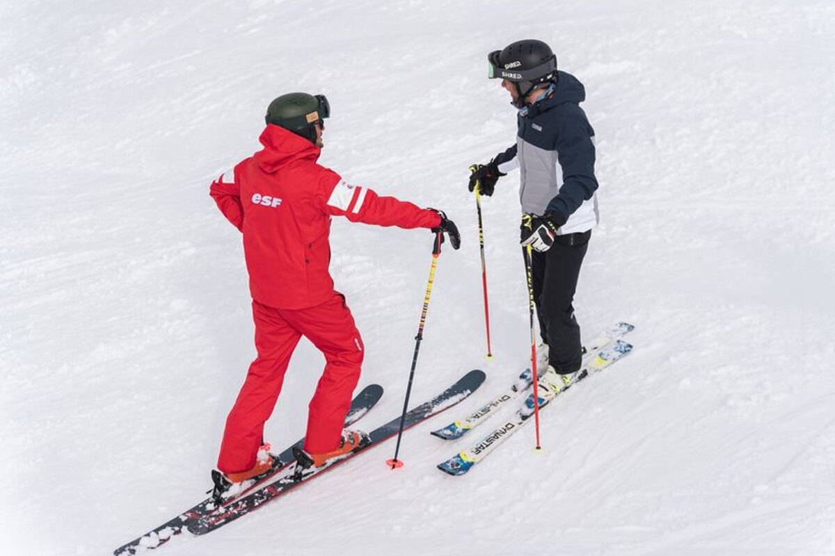 A man wearing a red esf jacket talks to another man on skis
