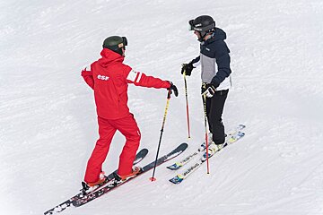 A man wearing a red esf jacket talks to another man on skis