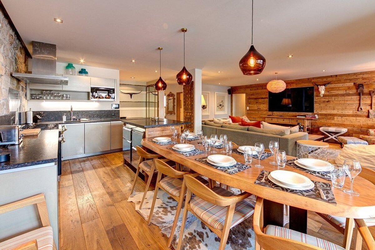 A chic, open-concept living space featuring a modern kitchen, a long wooden dining table set for a meal, and a cozy lounge with a rustic wood wall.