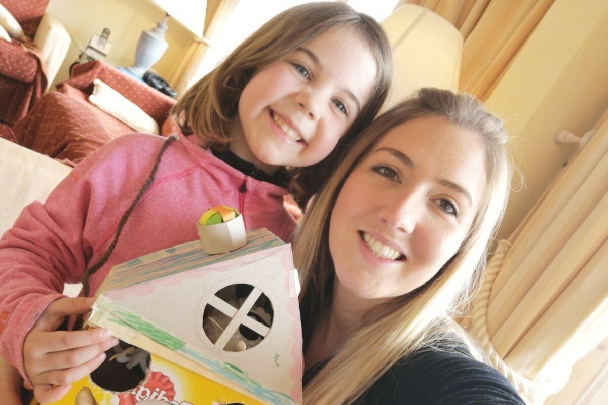 A smiling woman and a young girl joyfully hold up a colorful, homemade craft house, looking happy together in a cozy indoor setting.