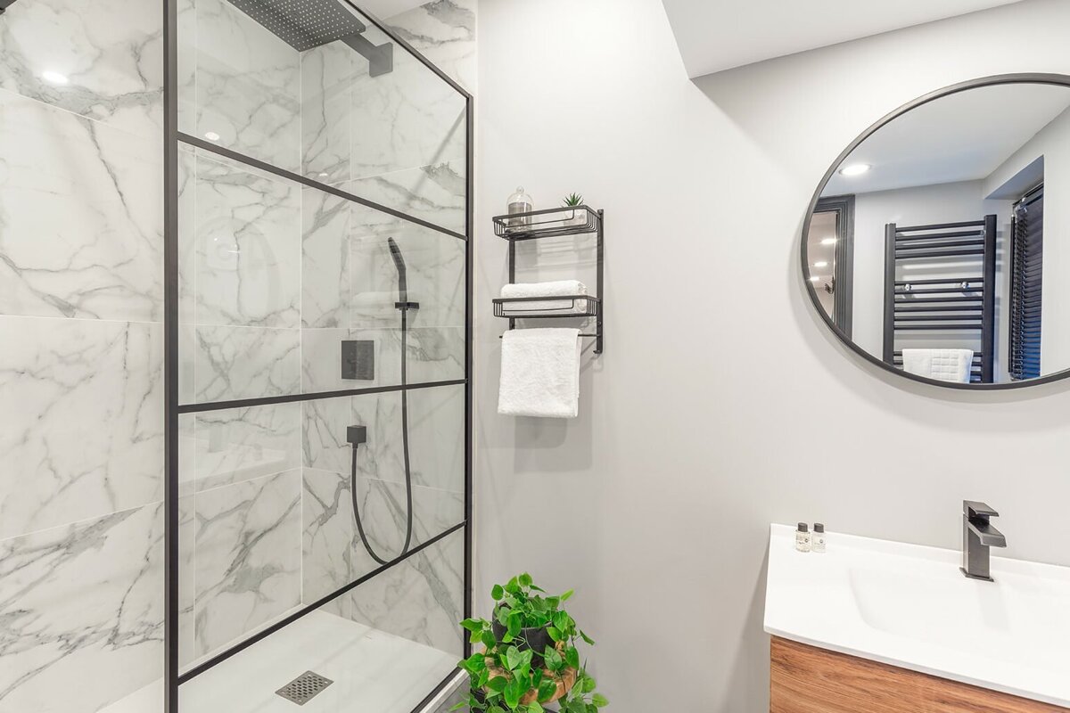A modern bathroom featuring a black-framed glass shower with marble-look tiles, a round mirror, floating wood vanity, and black fixtures.