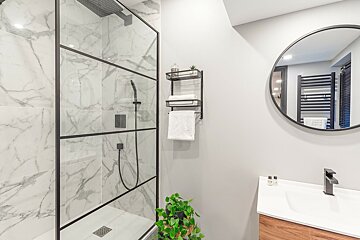 A modern bathroom featuring a black-framed glass shower with marble-look tiles, a round mirror, floating wood vanity, and black fixtures.