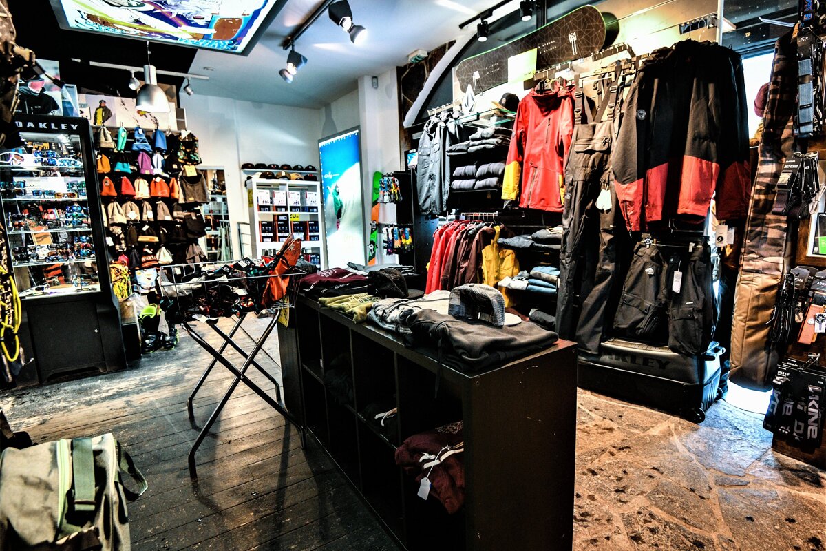 A brightly lit indoor store selling winter sports apparel and gear, including jackets, snowboards, goggles, beanies, and other outdoor clothing.