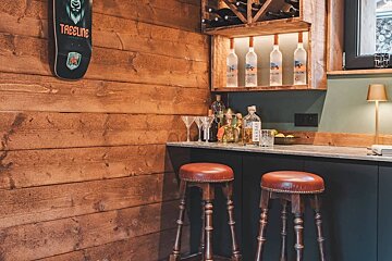 A rustic home bar with a wooden wall, illuminated liquor bottles, a marble counter, dark cabinets, two leather bar stools, and a skateboard.