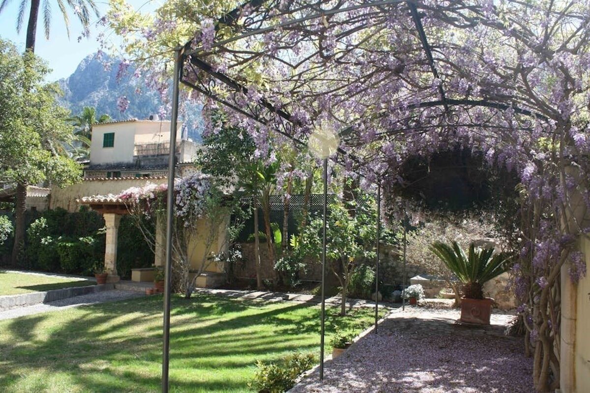 A vibrant garden path under a wisteria-draped pergola, with green lawns, a house, and a mountain in the background.