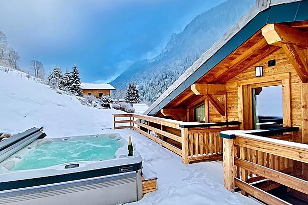 A cozy wooden chalet with a steaming hot tub on a snow-covered deck, overlooking a vast winter mountain landscape under a bright blue sky.