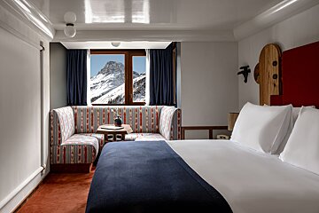 A modern bedroom with a breathtaking snowy mountain view. Features a cozy banquette, colorful patterns, a large bed, and sleek, warm decor.