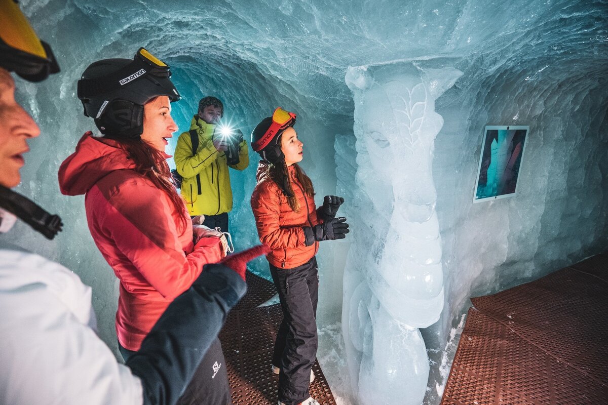 A group in winter gear explores an icy blue cave, admiring a sculpted ice horse and a framed picture on the frosty wall.