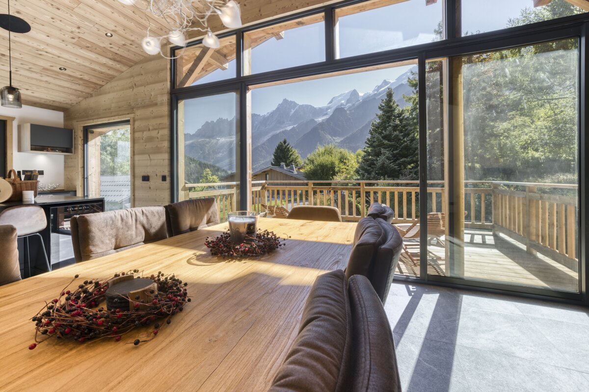 Modern wooden chalet dining room with panoramic mountain views through large windows and a balcony.