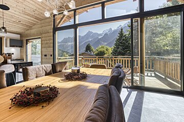 Modern wooden chalet dining room with panoramic mountain views through large windows and a balcony.