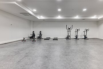 A minimalist, brightly lit gym features two rowing machines, two small treadmills, an elliptical, and two stationary bikes on a grey floor.