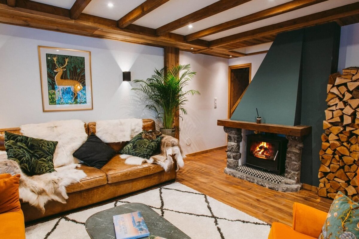 Cozy chalet-style living room with a crackling fireplace, stacked firewood, a leather couch, and wooden ceiling beams, creating a warm, inviting atmosphere.