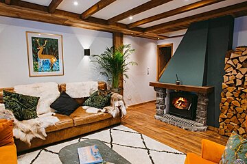 Cozy chalet-style living room with a crackling fireplace, stacked firewood, a leather couch, and wooden ceiling beams, creating a warm, inviting atmosphere.