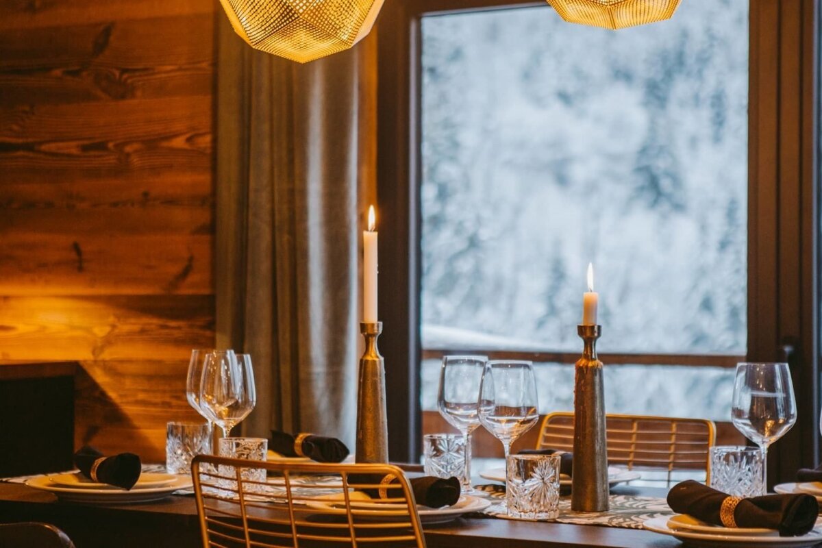 A cozy, candlelit dining table is set in a warm, wood-paneled room, with a snowy winter landscape visible through the large window.