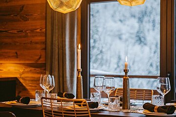 A cozy, candlelit dining table is set in a warm, wood-paneled room, with a snowy winter landscape visible through the large window.