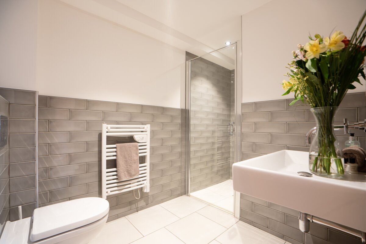A modern bathroom with grey subway tiles, white toilet, sink, towel warmer, glass shower, and fresh flowers on the counter.