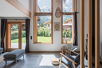 Bright chalet interior with large windows showcasing scenic mountain views, furnished with wooden accents and gray seating.