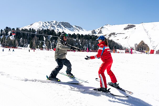 A ski instructor in red teaches a student to ski on a sunny, snowy mountain slope with distant peaks and pine trees.