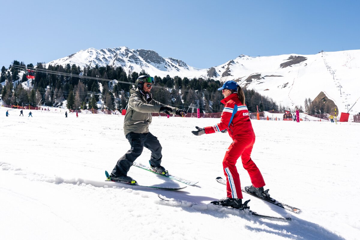 A ski instructor in red teaches a student in a green jacket on a sunny mountain slope with snowy peaks and trees in the background.