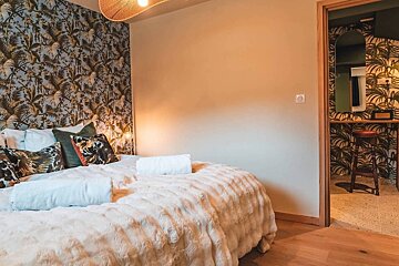 A tropical-themed bedroom features a fluffy bedspread against a leafy accent wall. An open doorway reveals an adjoining room with matching decor and a stool.