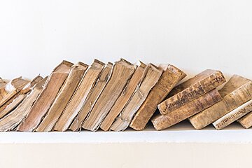 Worn, antique brown books lean and stack on a light surface against a white wall.