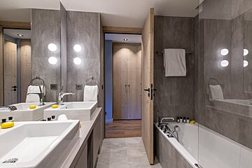 A modern bathroom features dual sinks, a bathtub, gray tiled walls, and wooden accents. Bright circular lights illuminate the space.