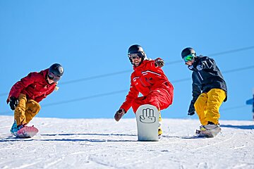 Three snowboarders are riding down a snowy slope and one has a hand on his board