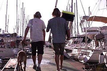 Two men and a dog are walking down a dock with boats in the background and one of the boats has the word gps on it